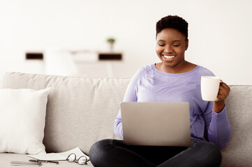 Smiling african american woman sitting on the couch, holding coffee mug and working on laptop. Lady...