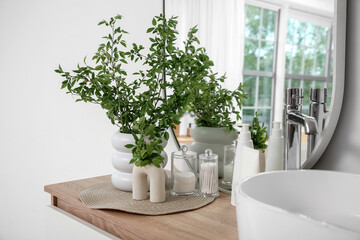 Vases of green blossoming branches with cosmetic products near sink on table in stylish bathroom