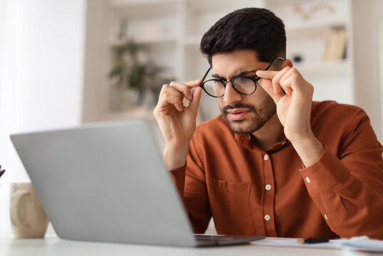 Error. Portrait of confused Arab guy sitting at desk using laptop, taking off glasses looking at pc screen and squinting. Worried man reading bad negative news, having poor eyesight problems