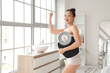 Happy young Asian woman with weight scales in bathroom. Diet concept