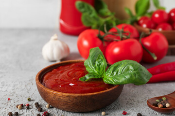 Wooden bowl of tasty ketchup with basil leaves, garlic and peppercorns on grey table