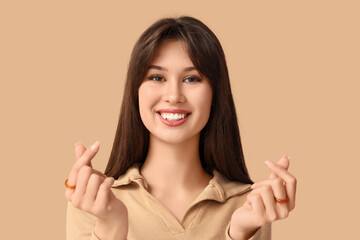 Smiling young woman making heart gesture on beige background, closeup