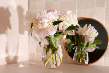 Mirror and vase with pink peony flowers on table near pink tile wall, closeup