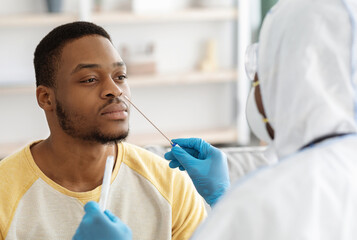 Black guy receiving nasal PCR test while staying home during COVID-19 pandemic, closeup. Unrecognizable doctor in protective suit taking nasal swab for young african american man patient