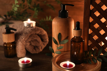 Composition with burning candles, green leaves and bottles of cosmetic products on wooden background, closeup