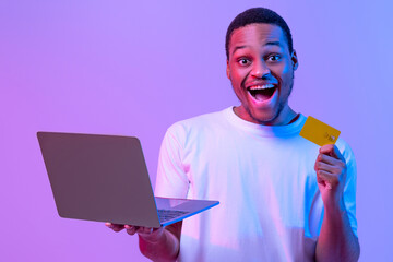 Online Shopping. Portrait of joyful black man with laptop and credit card standing in neon lighting...