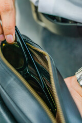 A man in a white shirt is putting his glasses into a black leather handbag.