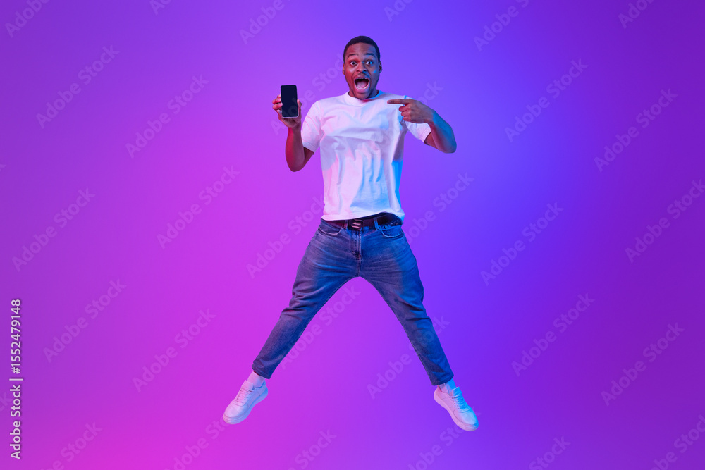 Poster portraif of amazed african man jumping over purple neon background and pointing at smartphone with b