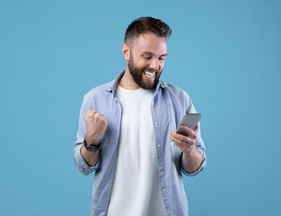 Portrait of happy bearded young man using smartphone, making winner gesture over blue studio background. Excited guy celebrating victory, achieving success, gesturing YES