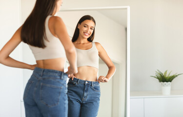 Cheerful young woman posing wearing jeans and smiling to her reflection in mirror after successful...