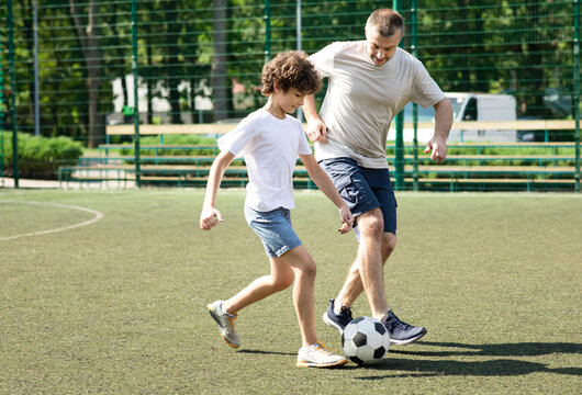 Active Lifestyle. Full length portrait of sportive father playing soccer with his son on green grass at stadium - Powered by Adobe