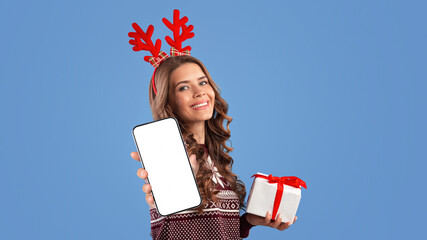 Lovely millennial lady in Xmas outfit holding gift box and pointing finger at camera on blue studio...