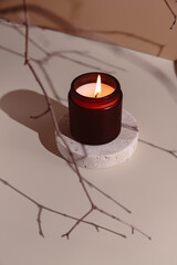 Minimalistic burning candle in a dark amber glass jar placed on a round travertine stone stand....