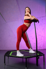 Energetic young fitness girl in red tracksuit posing standing on mobile mini trampoline. Jumping...