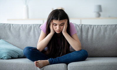 Covid related teen depression concept. Unhappy Indian teenage girl sitting on sofa at home with...