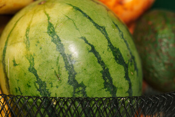 Close-up of a ripe watermelon resting in a black mesh fruit basket, surrounded by other fruits