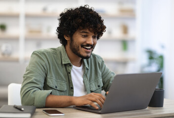 Remote job concept. Happy dark-skinned curly bearded young man in casual sitting at workdesk at...