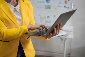 A middle-aged Asian woman sits at a work desk, deeply focused. Surrounded by charts and notes, she analyzes data, plans strategies, and prepares to lead a presentation or consultative session