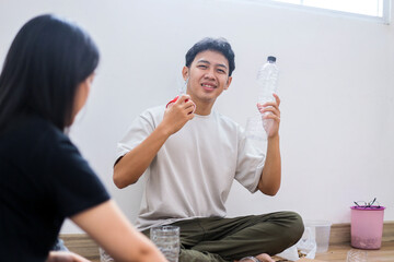 Fototapeta premium Smiling Young Asian Male Showing Scissors And Plastic Bottle While Sitting On Floor