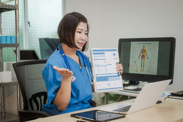 A middle-aged Asian female doctor sits alone at her desk in clinic, attentively reviewing patient...