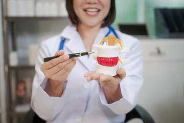 A middle-aged Asian female doctor sits at her desk, pointing to a spinal model with a highlighted red section. She explains spinal conditions like herniated discs or nerve compression to a patient
