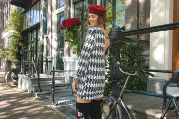 Beautiful elegant  woman in black and white knit dress, showing open back of the dress and  holding red roses in front of bicycle on streets of Amsterdam city - Tourist enjoying summer vacation 