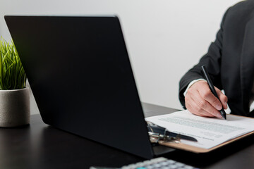 Businessman reviewing and signing contract on clipboard at desk with laptop and calculator
