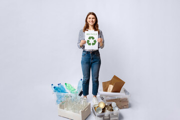 A woman stands confidently in a studio, holding a recycling sign while surrounded by various...