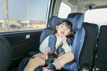 A 6-month-old baby and a 3-year-old Japanese girl sit in car seats in the back seat, gazing forward...