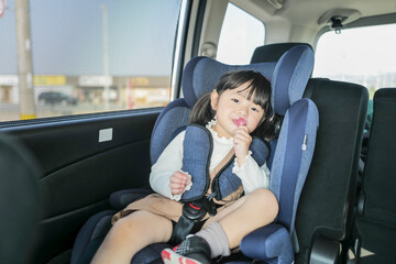 A 6-month-old baby and a 3-year-old Japanese girl sit in car seats in the back seat, gazing forward...