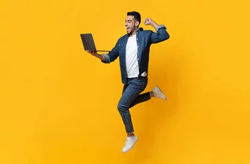 Gordijnen Emotional arab young man jumping up, using new modern laptop, raising fist up and smiling over yellow studio background, trading online, copy space for text or advertisement, panorama © Prostock-studio