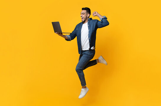 Fototapeta Emotional arab young man jumping up, using new modern laptop, raising fist up and smiling over yellow studio background, trading online, copy space for text or advertisement, panorama