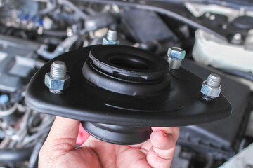 Top Strut Mount, A close-up of hand holds a black automotive strut mount with visible bolts over a car engine.