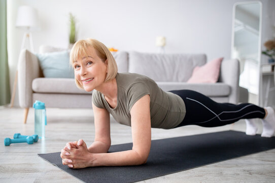 Home training concept. Smiling senior woman doing elbow plank on yoga mat in living room. Positive mature lady doing her workout routine, exercising indoors, strengthening her core muscles - Powered by Adobe
