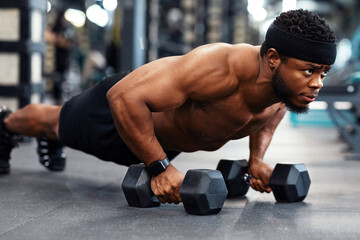 Concentrated black guy sportsman pushing up with dumbbells. Muscular african american bodybuilder shirtless man holding barbells and making push-ups from floor, work out at gym