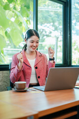A joyful young woman in a pink blazer celebrates success while working on her laptop in a bright,...