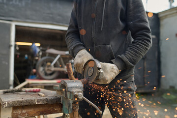 Middle aged Caucasian man using angle grinder to cut metal part while assembling custom bike in workshop, wearing protective gloves, sparks flying from cutting process, garage background visible
