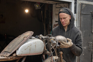 Caucasian young adult man repairing custom motorcycle in workshop, wearing gloves and hoodie,...