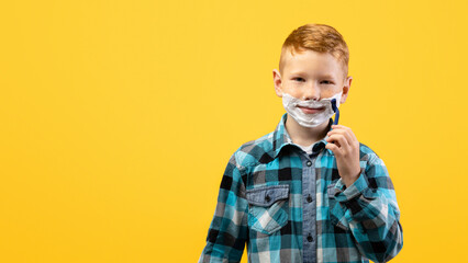 Playful little young boy shaving face over yellow studio background, imitating his dad, copy space. Cute redhead kid with shaving foam on his face holding razor, acting like adult man
