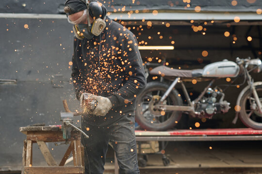 Middle aged man wearing protective gear using angle grinder while repairing custom motorcycle in workshop, sparks flying as he works on metal part near vintage bike
