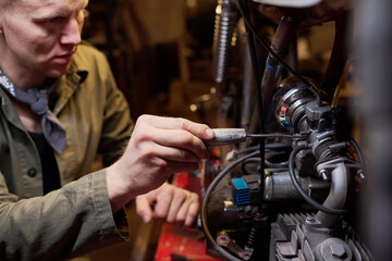 Obraz premium Caucasian young adult man repairing custom motorcycle in workshop, focusing on engine components with hand tools, working attentively in garage setting, partial face visible
