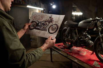 Caucasian middle aged man holding motorcycle blueprint while assembling and repairing custom bike in workshop, focusing on design details and mechanical components on workbench