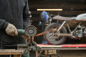 Caucasian middle aged man using angle grinder to cut metal pipe while assembling custom motorcycle in workshop, sparks flying as bike frame visible in background, wearing protective gloves