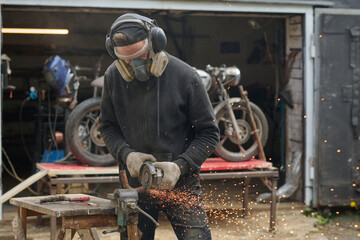 Caucasian middle aged man wearing protective gear using angle grinder while repairing custom motorcycle in workshop garage, sparks flying as he works on metal component