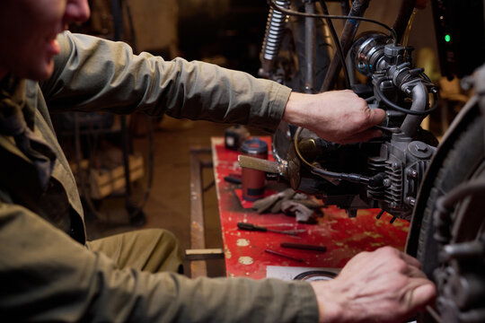 Caucasian young adult man assembling and repairing custom motorcycle engine in workshop, focusing on mechanical components with hands working on intricate parts at cluttered workbench