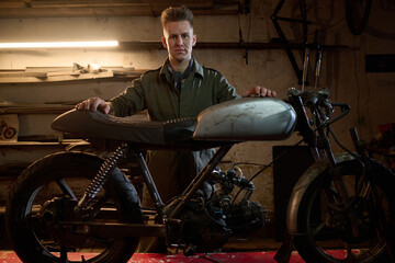 Caucasian young adult man standing behind custom motorcycle assembling and repairing bike in workshop or garage, focused expression, hands resting on seat and handlebars