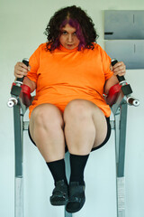 Beautiful overweight Latina woman wearing colorful 80s-style clothing and makeup, exercising with abdominal muscles on a machine in a gym, very positive. Effort, improvement, and overcoming.