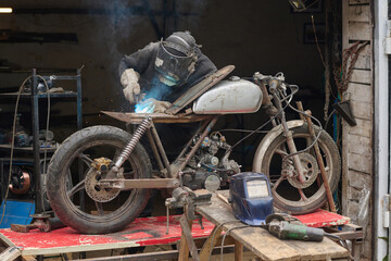 Middle aged Caucasian man welding custom motorcycle frame in workshop, wearing protective helmet and gloves, focusing on assembling and repairing bike on workbench