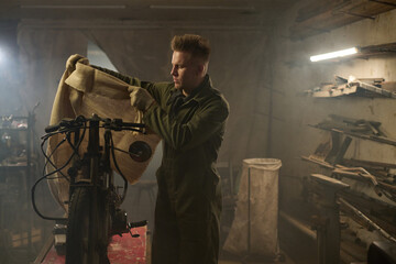 Caucasian young adult man assembling custom bike in workshop, wearing work gloves and coveralls, standing at workbench and handling parts, surrounded by tools and equipment