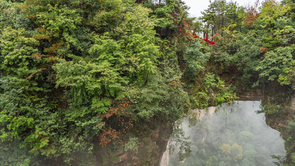 A natural stone bridge between the rocks. The arch over the gorge. There is green vegetation on the steep slopes of the mountains. Red ribbons on the railing of the footpath. Fog. China. Zhangjiajie